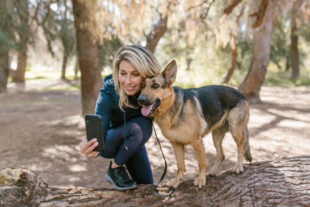 Smiling woman taking a selfie with her German Shepherd dog in a sunny forest setting.